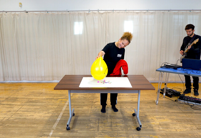 Person in a dance studio holding a yellow balloon upright on a table. Behind them is a person with an electric guitar.