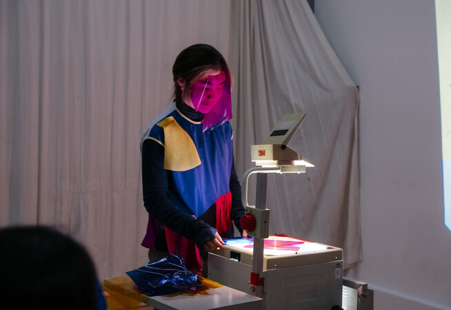 A performer stands over an overheard manual projector with a pink transparent gel masked over their face, projecting pink onto a white wall. They are wearing a black blue red and yellow colour blocked top, the wall behind them is covered with off white draped fabric.