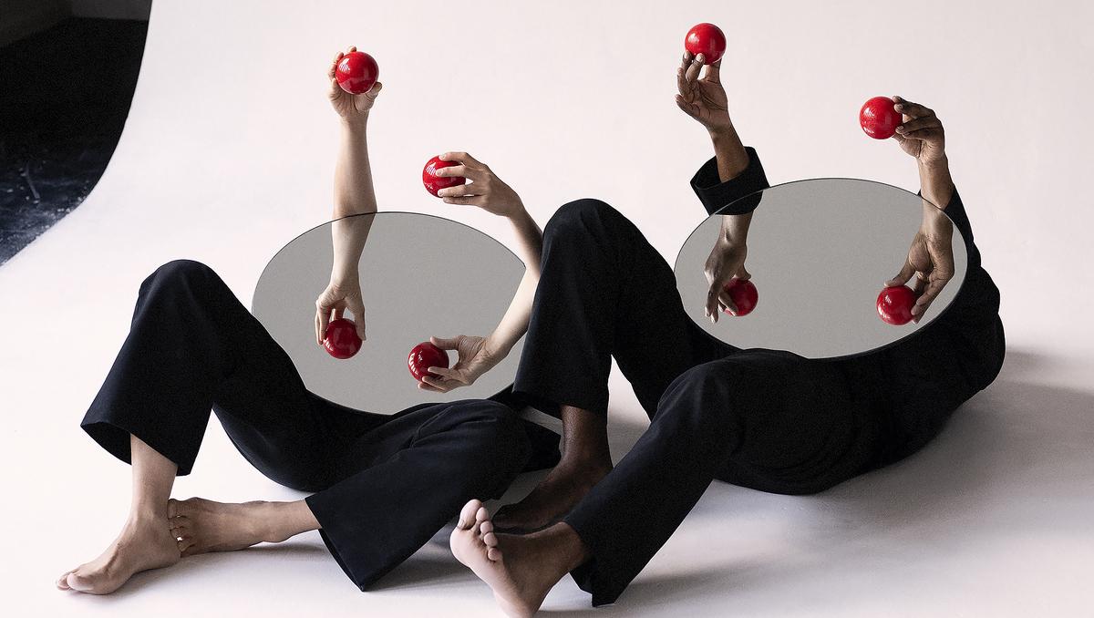 A surreal photo of two figures dressed in black lying against a white background with their heads obscured by mirrors, holding red juggling balls