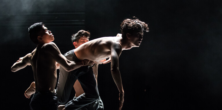 Three dancers: two with their hands clasped, lifting up the third. Dark backdrop and dramatic lighting.