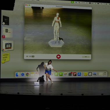 A performer moves in front of a giant projection screen, blurring the boundary between reality and the digital world. The scene prompts a reconsideration of the body through duality and reflection.