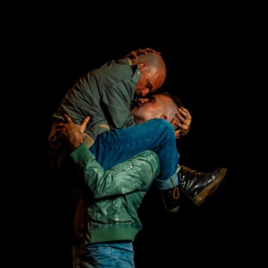 Two male dancers embracing against a black background.