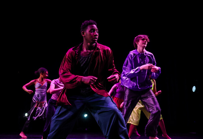 A group of young dancers between 14 and 19 on a stage against a black background. They are wearing satin clothing in a mixture of rich colours.