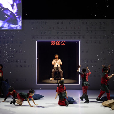 A photo of a cohort of child performers dancing on stage with an adult drummer sat in the background with a motion sensor drumkit and headphones. There is also a projection of a panda in the background.
