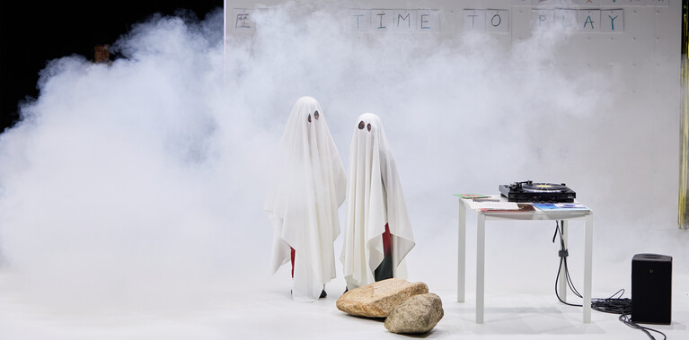 A photo of two child performers dressed as ghosts on a stage, wearing white sheets. A record player and stage smoke surrounds them.