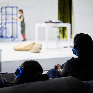 A photo of the backs of audience members, with headphones on, watching child performers on stage.