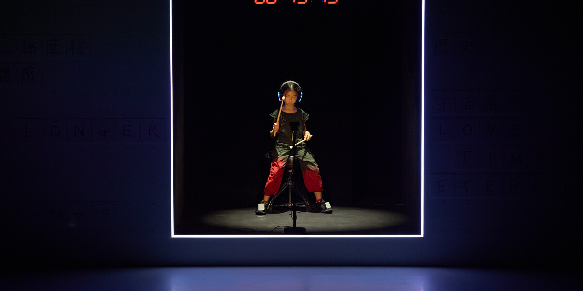 A photo of a child performer sat in a box with headphones on playing a motion sensor drumkit. There is a red digital clock above them.