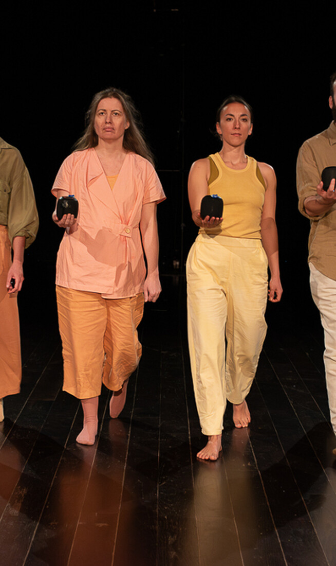 Four performers wearing soft pastel tones walk purposefully towards the camera in unison, each with one arm extended in front of them, portable bluetooth speakers in the palm of their hands.