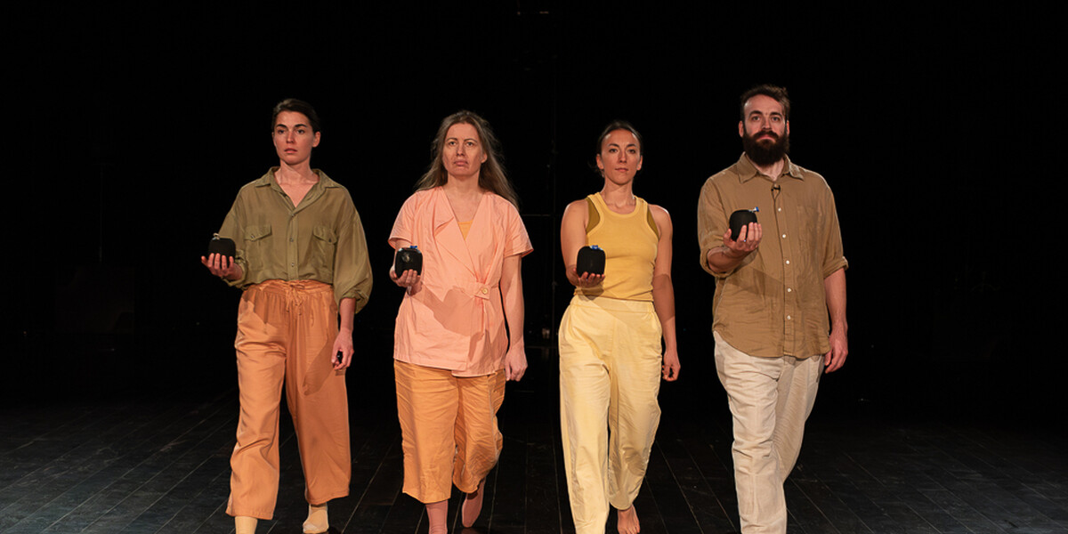 Four performers wearing soft pastel tones walk purposefully towards the camera in unison, each with one arm extended in front of them, portable bluetooth speakers in the palm of their hands.