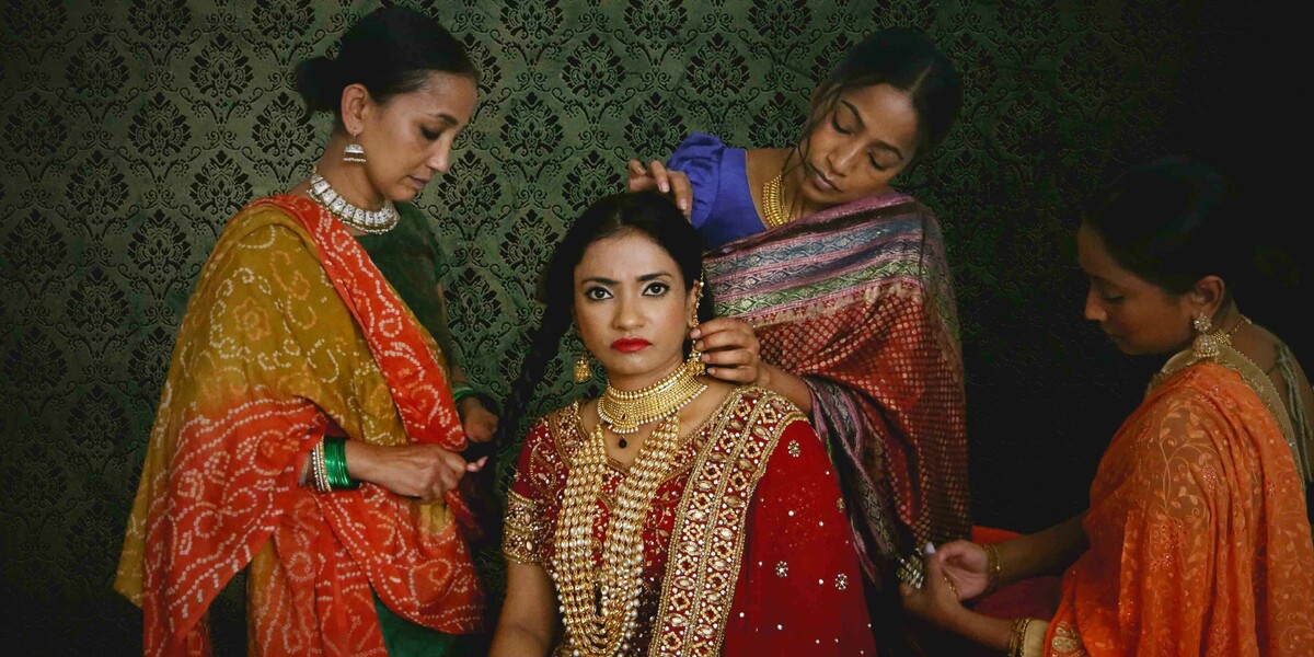 A woman wearing a sahri is looking down the camera and having her hair done by three other woman who are also wearing sahris. There is a decorative green wallpaper in the background.