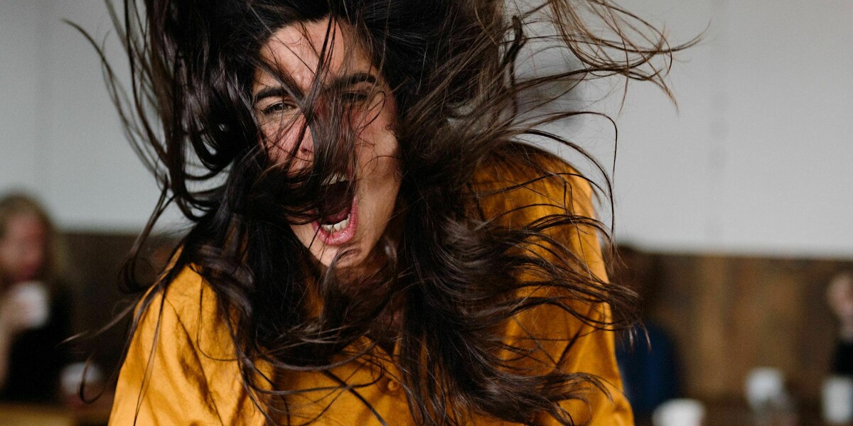 A woman wearing an orange shirt bangs her fist angrily on the table.