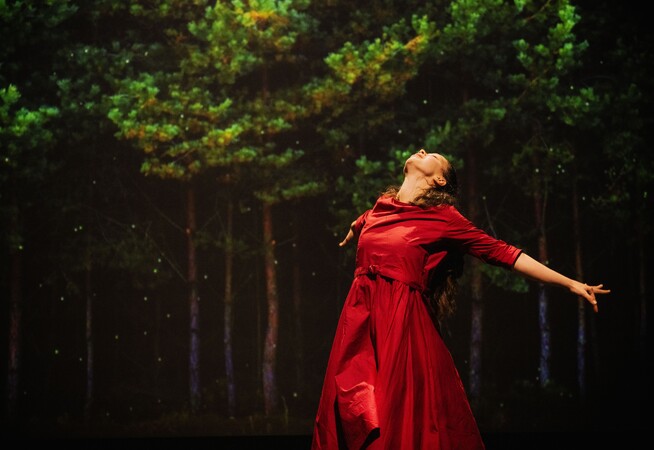 A woman wearing a red dress leaning backwards. Trees are on a screen in the background behind her.
