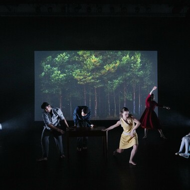Six performers in motion on a dark stage, with the stage walls visible. Behind them is a projection of trees at night