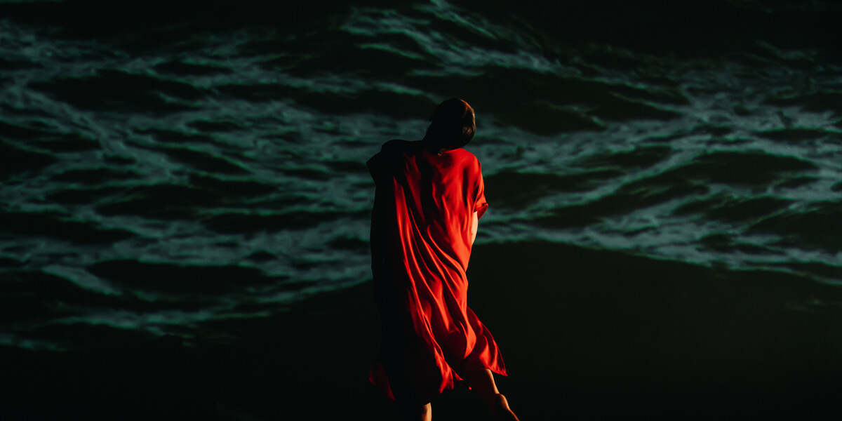 A figure draped in a red cloth, facing away from the camera towards the dark outline of a natural landscape in the background.