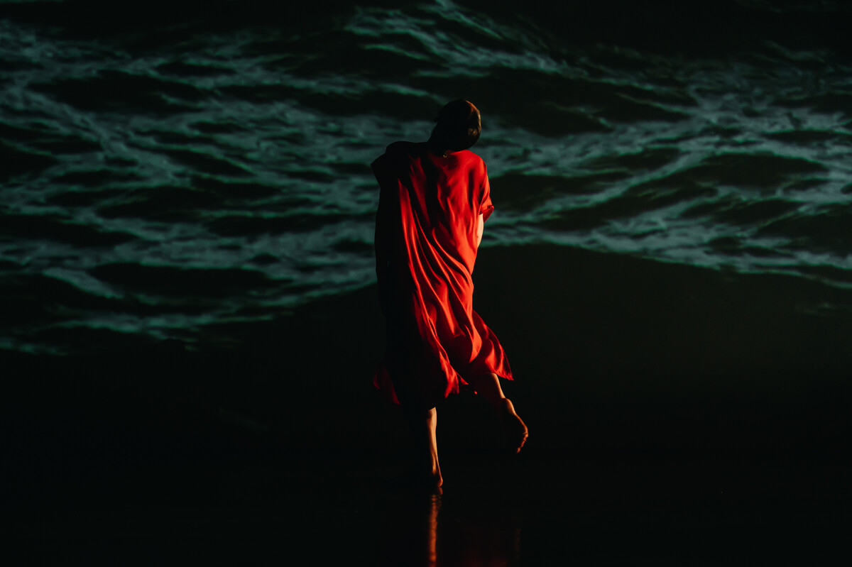 A figure draped in a red cloth, facing away from the camera towards the dark outline of a natural landscape in the background.