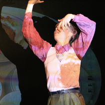 A dancer’s upper body wearing a rose-coloured vest over a pink shirt with dark green plaid trousers in the style of the 60s. The dancer has their hands in the air and images of china plates are projected over them and the wall behind.