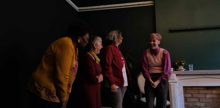 A dark green setting which suggests a living room. Three older women* stand next to one younger person. They look at each other and smile or laugh.