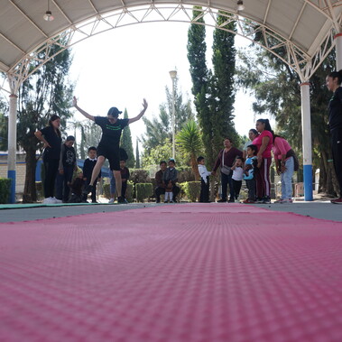 A red rubber mat in a school playground. Preschool children and teachers observe as a dance teacher jumps.
