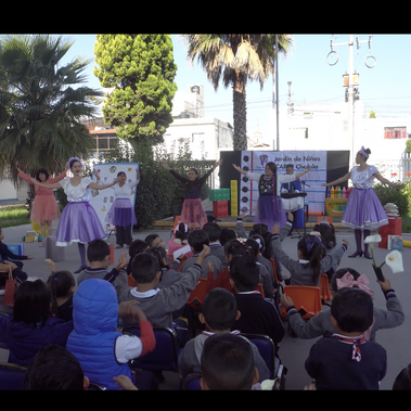 A school assembly outside. Six dancers perform for school children; arms raised in the air. Rows of children watch from seats, some also raising their hands.