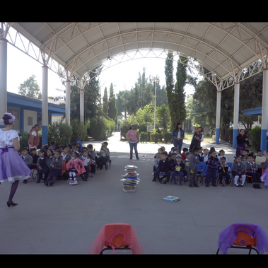 Two dancers in purple skirts perform for an audience of children seated on chairs in the school yard. A pile of books is in between the dancers.