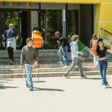 Young teens, ages 12-15, carrying colourful chairs on the steps in front of their school on a sunny day.