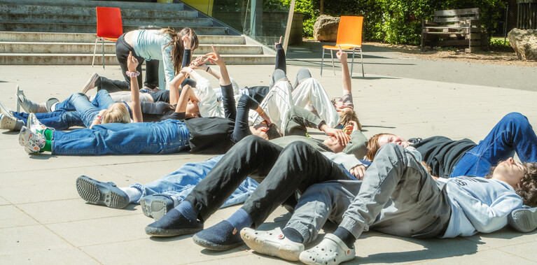 Young teens lying on the ground, gazing and pointing at the sky in front of their school on a bright day.