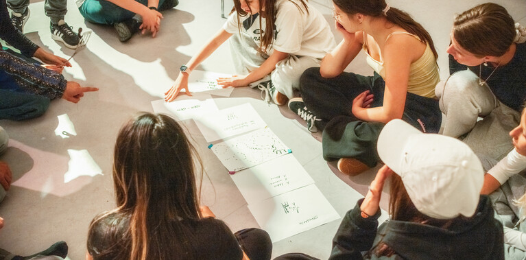 A group of young teens, sitting in a circle on the floor, discussing notes and drawings, with one girl explaining.