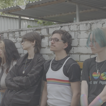 A group of people standing in a row leaning against a long wall, looking ahead into the distance. Some have their eyes closed some open.