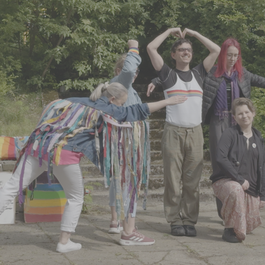 A group of people make a formation standing closely with their bodies in a colourful outdoor setting, surrounded by LGBTQ+ pride signs and rainbow-themed decorations.