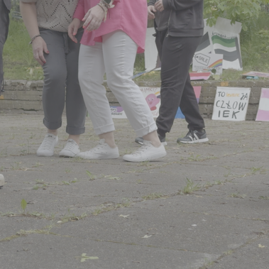 Close-up of people’s legs and feet standing on a paved area, some wearing rainbow socks and pride-themed signs visible in the background.