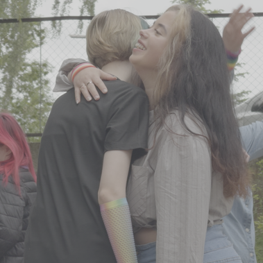 Two people share a joyful hug at an outdoor gathering, surrounded by others celebrating with smiles and softness.