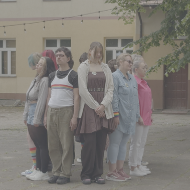 A group of people stands back-to-back in a circle facing outward in an outdoor courtyard, wearing pride-themed clothing and accessories.