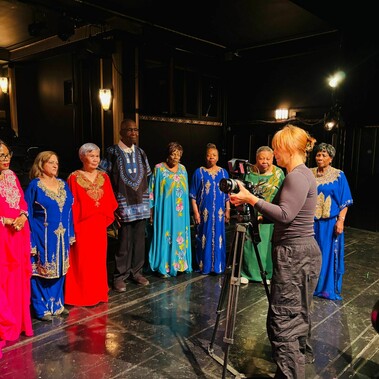 Project participants in a room posing for a photo shoot, wearing glamorous gowns in various bright colours. A camerawoman stands before them holding a camera.