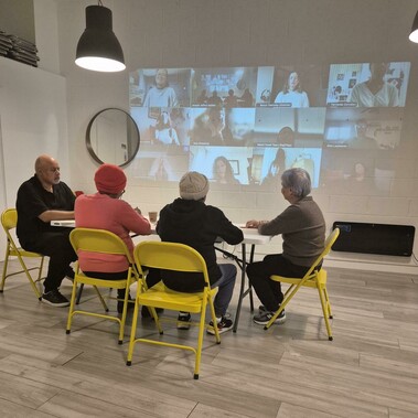 Four adults sit on yellow chairs at a table before an image of many students beamed onto the wall. The live and video participants are looking at each other.