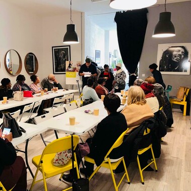 A room with tables and chairs in a U shape. Many adults sit with drinks and snacks before them, listening attentively to a man who is reading from papers at the front of the room.