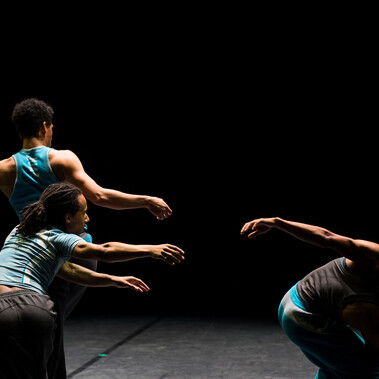 Three Black men dance on a dark stage in a triangle formation, wearing blue, their arms reaching for one another