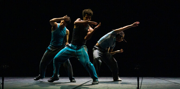Three Black men dance on a dark stage in a triangle formation, wearing blue