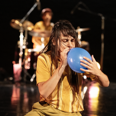 A dancer with long dark hair kneels and blows into a blue balloon, with a drummer visible in the blurred background.