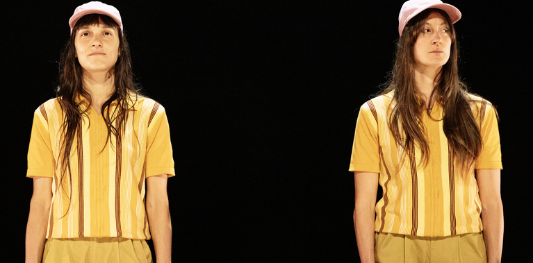 Two almost identical dancers with long dark hair wearing matching yellow striped shirts, tan shorts, and pink hats stand side-by-side, the performer on the left has a soft almost smile while the one on a right a more mischievous look. They stand together against  against a black background.