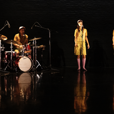 A drummer sits at a red drum kit on the left side of a dark, reflective stage. On the right, two performers stand with microphones, all three in matching outfits, yellow tops and bottoms as well as pink knee-high socks.