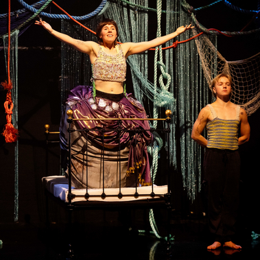 Two performers are on a stage with a set resembling a tangled net of ropes. One performer sits atop a bed frame with their arms outstretched, wearing a large, purple costume. The second performer stands beside the bed, looking to the side.