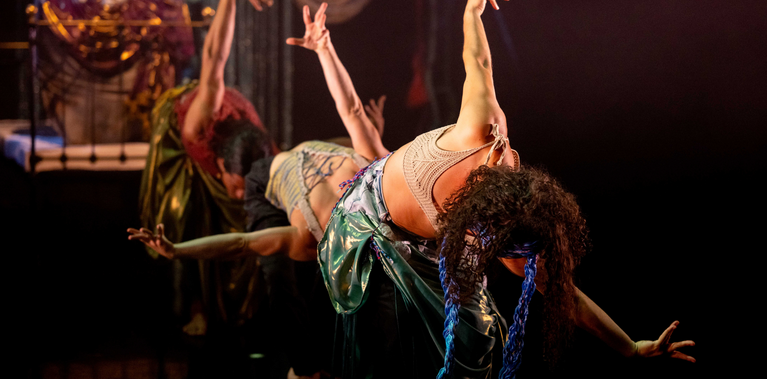 Three performers are on a dark stage, leaning back in a curtsy position with their arms raised. The central performer, in a shiny green skirt and a light top, is the clearest, while the others are slightly blurred in the foreground, creating a synchronized, fluid motion