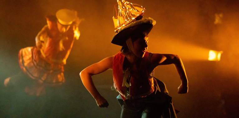 A performer in a captain's hat with a lit-up model ship on top crouches forward on a dimly lit, foggy stage. The scene is cast in an orange glow from the stage lights.