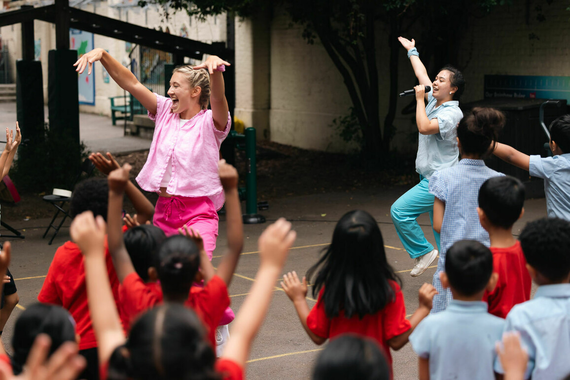 Two dancers - one in pink and one in blue - dance energetically in a school playground whilst children wave their arms and jump in front of them