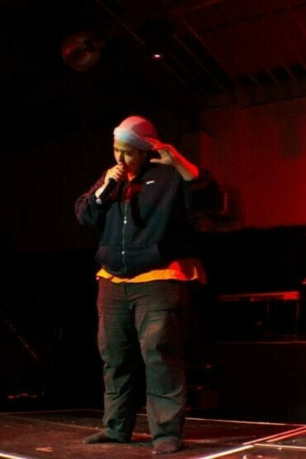 A person wearing a headscarf, hoodie and trousers stands onstage, speaking intently into a microphone.