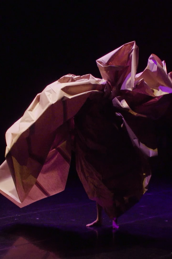 A person stands barefoot on stage, their body from the calves up entirely covered by a large, crumpled sheet of paper.