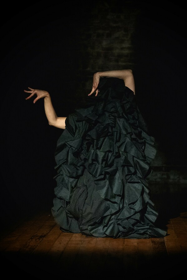 A person on stage turned away from the audience, wearing a flamboyant, ruffled costume and raising their arms into an angular shape.