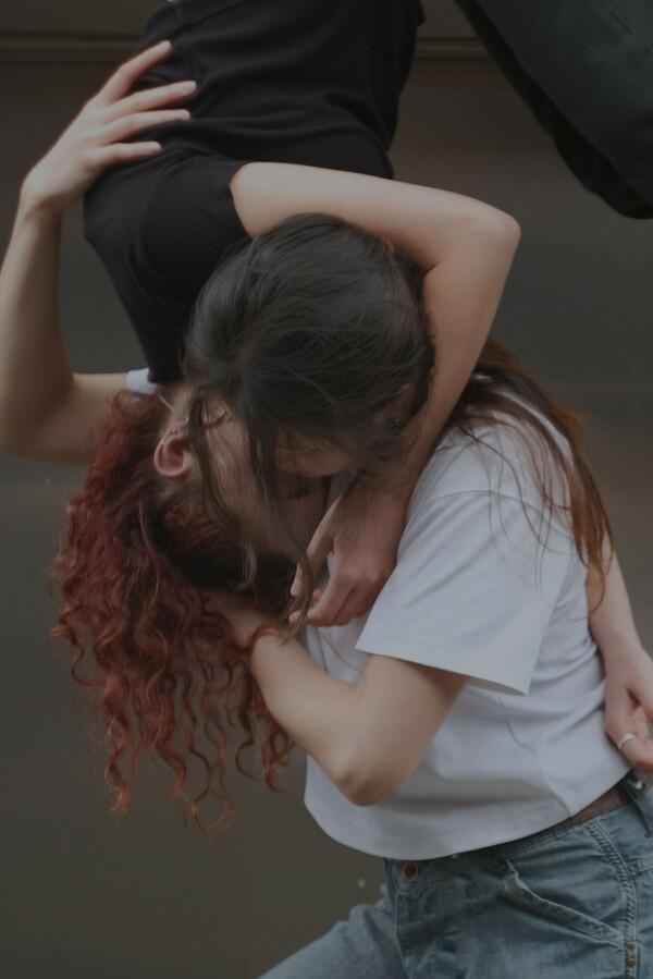 Two people are embracing, with one suspended above and hanging downward, wearing a black T-shirt and jeans, while the other stands below, wearing a white T-shirt and blue jeans.