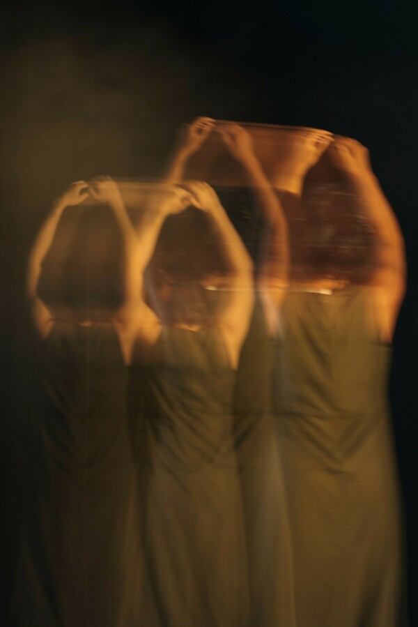 A blurred photo shows a group in uniform grey dresses standing in formation with their backs to the audience, arms raised overhead, while one figure stands off to the right, set apart with arms resting by her sides.