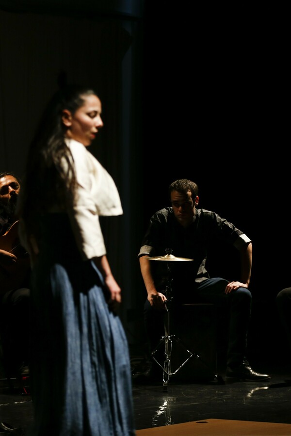 Woman standing on stage in front of a group of musicians, wearing a long dark skirt and white jacket, captured in between movements with her arms by her sides.